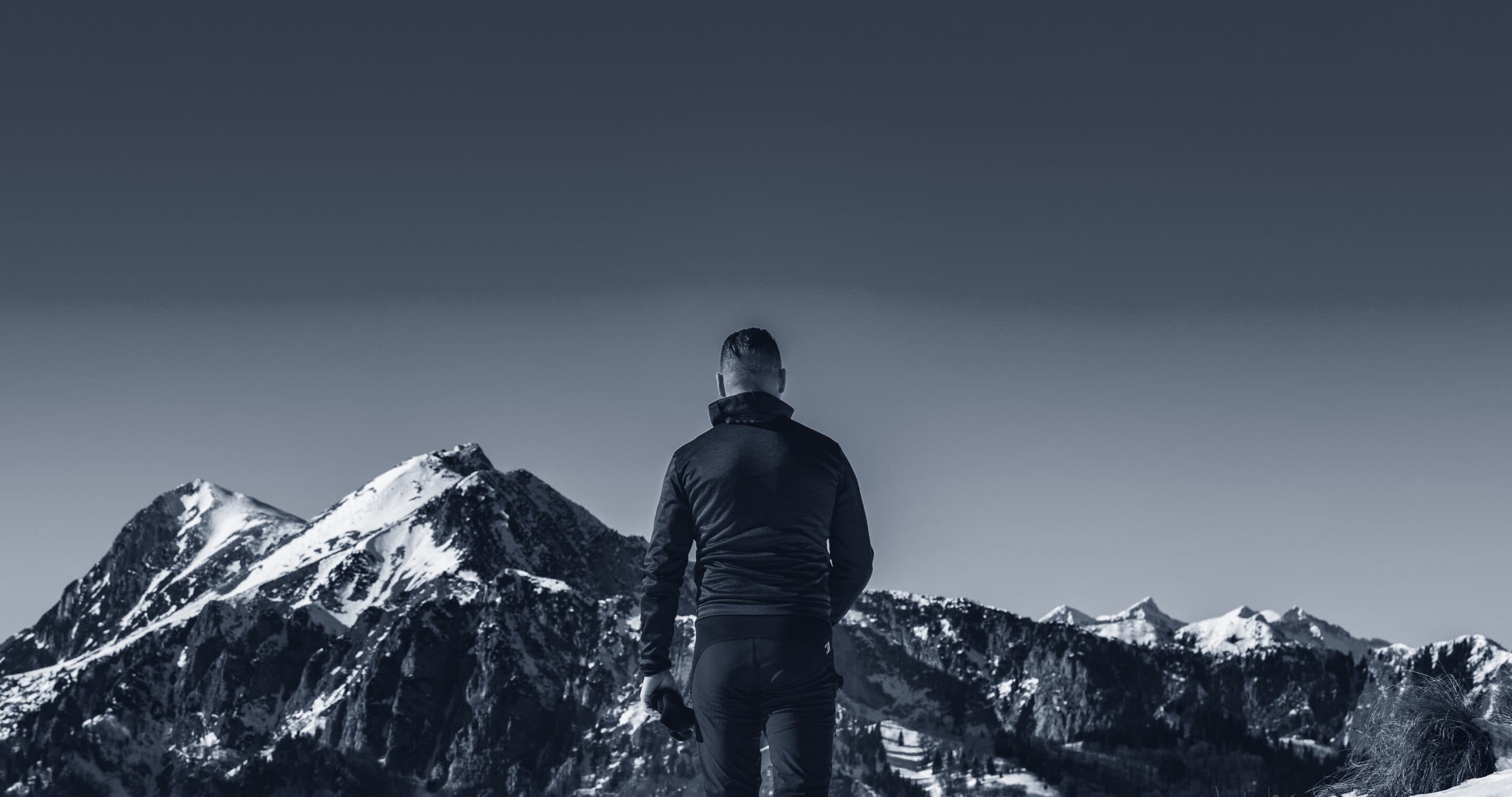 A man in a dark jacket standing on a snowy ridge, looking towards snow-capped peaks